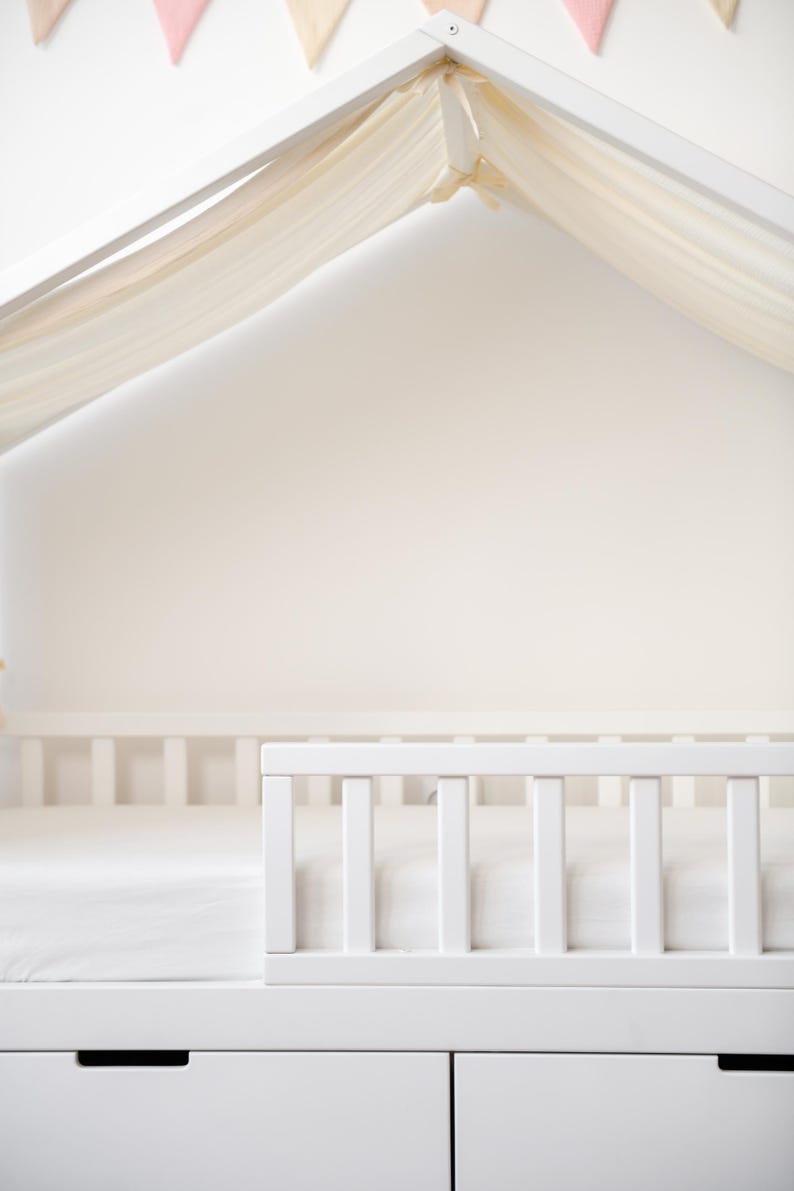 May include: A white wooden house-shaped bed frame with a white mattress and a white railing. The bed has two drawers at the bottom. A sheer, cream-coloured fabric drapes over the top of the frame. Pink and beige pennant flags are visible.