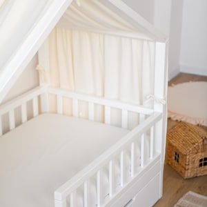 May include: A white wooden bed frame in the shape of a house, featuring a canopy and a white mattress. The bed has a slatted side rail and a drawer underneath. Cream-coloured fabric drapes from the canopy. A small woven house-shaped basket is nearby.