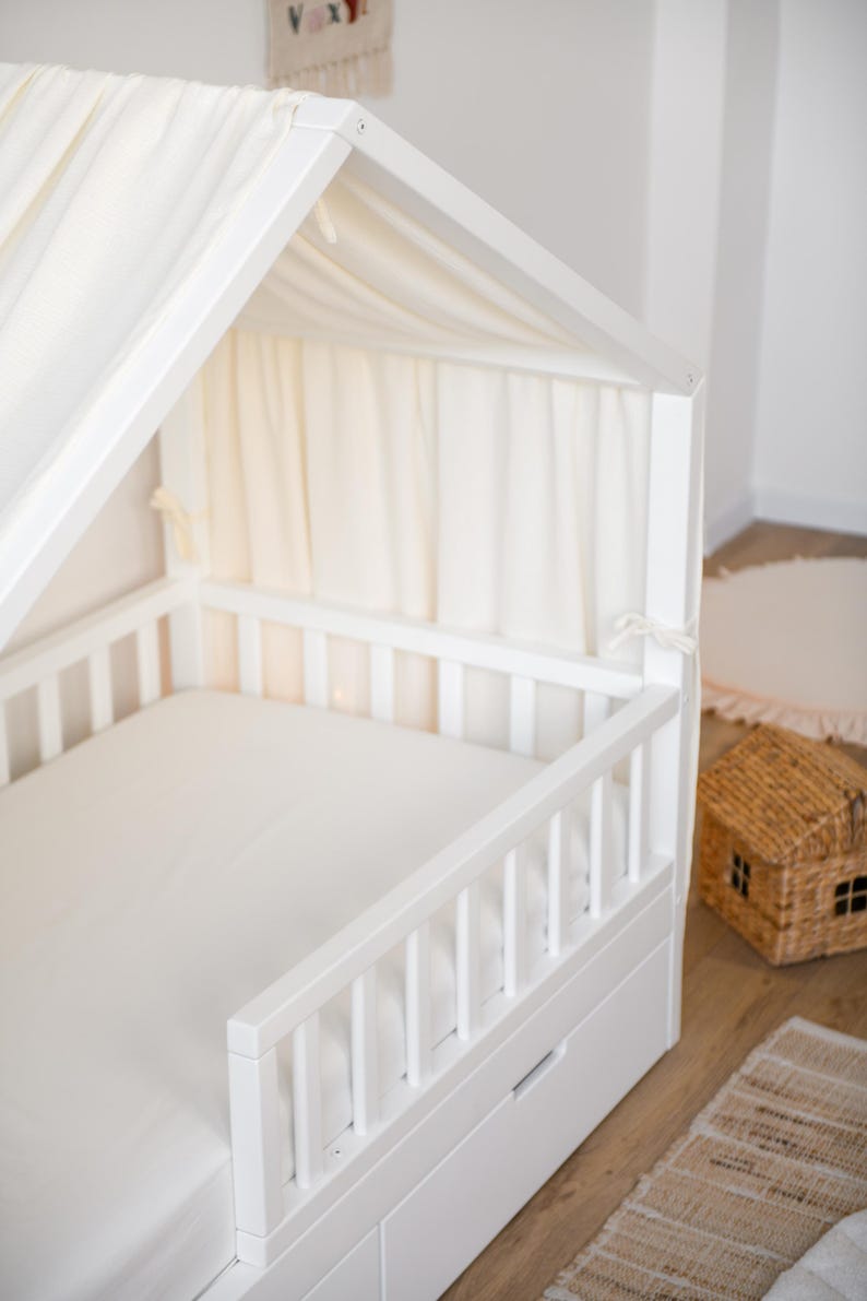 May include: A white wooden bed frame in the shape of a house, featuring a canopy and a white mattress. The bed has a slatted side rail and a drawer underneath. Cream-coloured fabric drapes from the canopy. A small woven house-shaped basket is nearby.