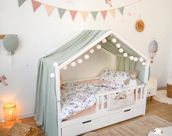 May include: A white wooden house-shaped bed with a green canopy and string lights. The bed has a floral patterned comforter and a teddy bear. The room is decorated with wall decals of hot air balloons and airplanes, and a garland.