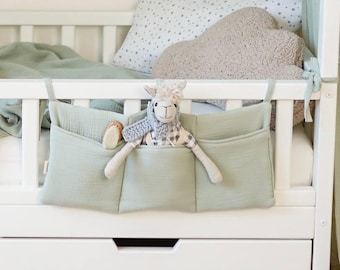 May include: A white crib with a sage green organizer hanging on the side. The organizer has three pockets and a stuffed llama toy inside. A cloud-shaped pillow and a star-patterned pillow are on the bed.