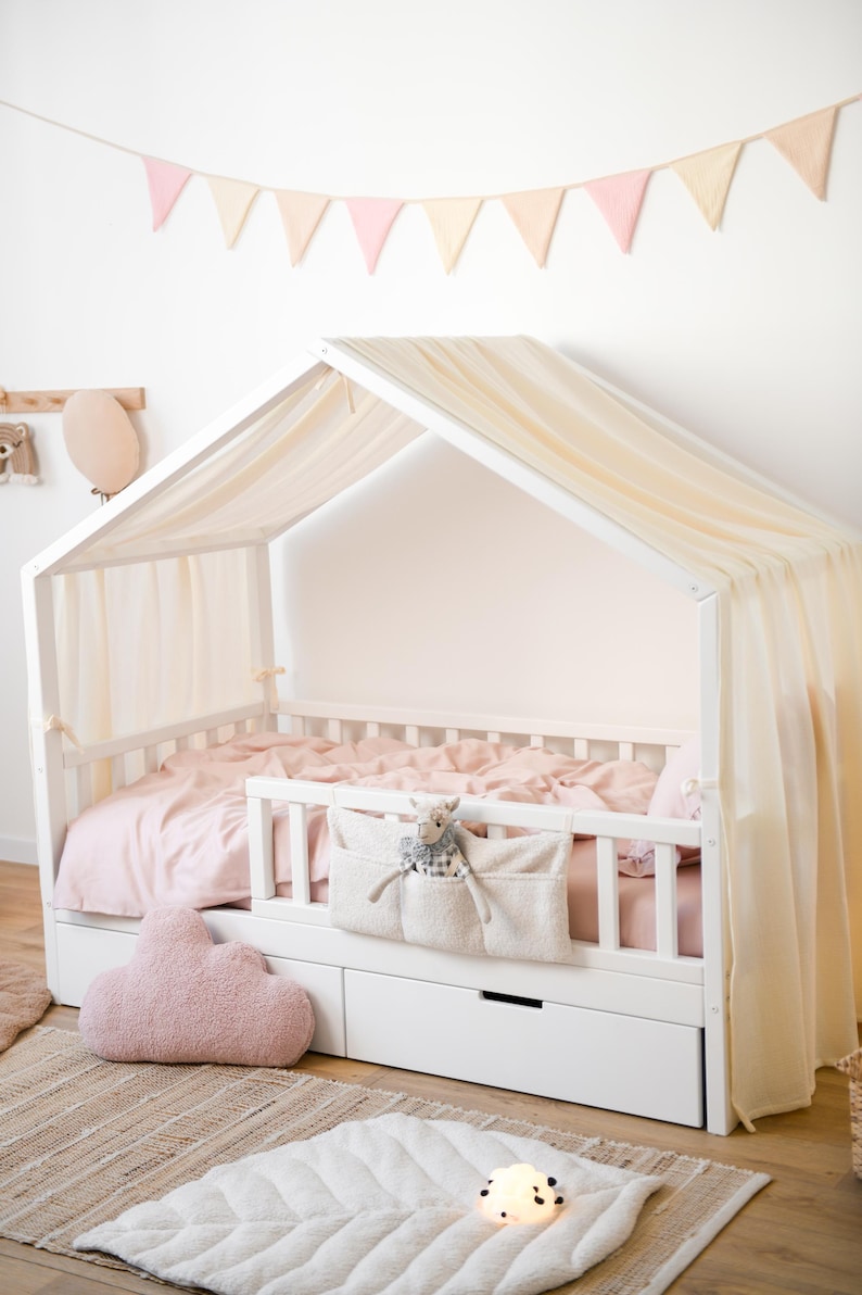 May include: A white house-shaped bed with a canopy and pink bedding. The bed has a storage drawer and a fabric organiser with a stuffed animal. A cloud-shaped pillow and a leaf-shaped rug are on the floor.