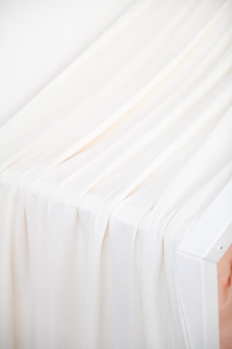May include: Cream-coloured fabric draped over a white structure, forming gentle folds and shadows. The fabric looks lightweight and flowing, with a delicate texture. The overall aesthetic is minimalist and elegant, suggesting a focus on simplicity and clean lines.
