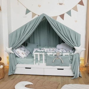 Op de afbeelding: Een wit houten bedframe in de vorm van een huis met twee opberglades eronder. Het bed is gedrapeerd met saliegroene stoffen gordijnen, voorzien van witte stervormige kussens aan de zijkanten en een kleine knuffel in een hangend vak.