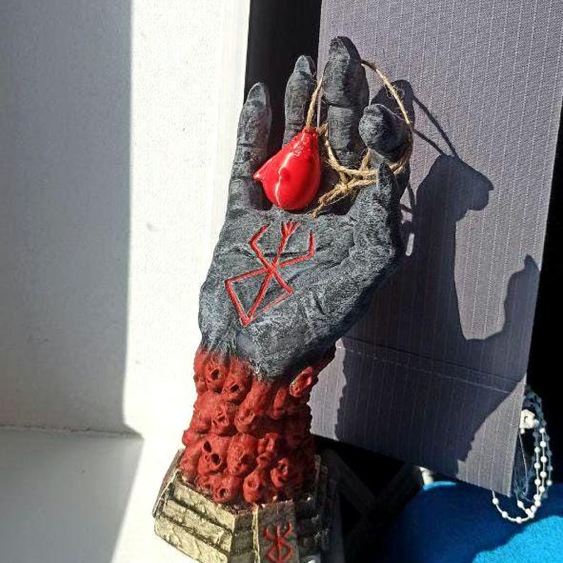 Puede incluir: Una escultura de mano negra con un colgante de coraz&oacute;n rojo y un s&iacute;mbolo rojo en la palma. La mano est&aacute; rodeada por una base de cr&aacute;neos rojos.
