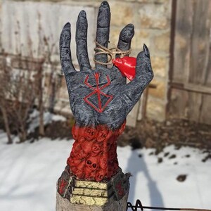 Puede incluir: Una escultura de mano negra con un s&iacute;mbolo rojo en la palma, rodeada por un anillo de caras rojas. La mano sostiene una peque&ntilde;a campana roja.