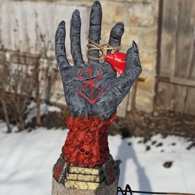 Puede incluir: Una escultura de mano negra con un s&iacute;mbolo rojo en la palma, rodeada por un anillo de caras rojas. La mano sostiene una peque&ntilde;a campana roja.