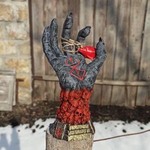Puede incluir: Una escultura de mano negra con un coraz&oacute;n rojo en la palma. La mano est&aacute; cubierta con un patr&oacute;n de caras rojas y negras. La mano sostiene un coraz&oacute;n rojo en la palma. La mano est&aacute; sobre una base de madera.