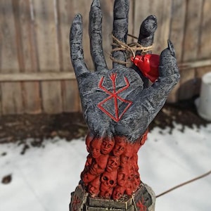 Puede incluir: Una escultura de mano negra con un s&iacute;mbolo rojo en la palma. La mano sostiene un peque&ntilde;o coraz&oacute;n rojo. La mano est&aacute; rodeada por una base de caras y cr&aacute;neos rojos.