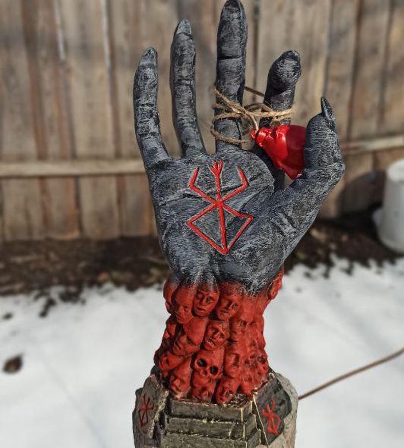 Puede incluir: Una escultura de mano negra con un s&iacute;mbolo rojo en la palma. La mano sostiene un peque&ntilde;o coraz&oacute;n rojo. La mano est&aacute; rodeada por una base de caras y cr&aacute;neos rojos.