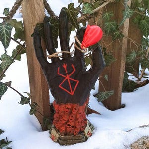 Puede incluir: Una escultura de mano negra con un s&iacute;mbolo rojo en la palma, rodeada por un anillo de peque&ntilde;as caras rojas que parecen humanas. La mano sostiene un globo rojo.