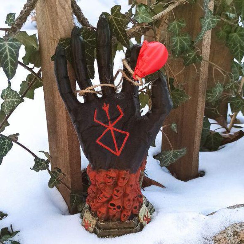 Puede incluir: Una escultura de mano negra con un s&iacute;mbolo rojo en la palma, rodeada por un anillo de peque&ntilde;as caras rojas que parecen humanas. La mano sostiene un globo rojo.
