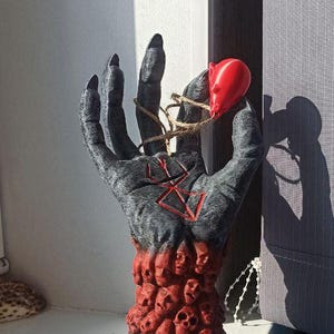 Puede incluir: Una escultura de mano negra con un s&iacute;mbolo rojo en la palma. La mano sostiene un objeto rojo con una cuerda atada. La mano est&aacute; hecha de muchas caras peque&ntilde;as.