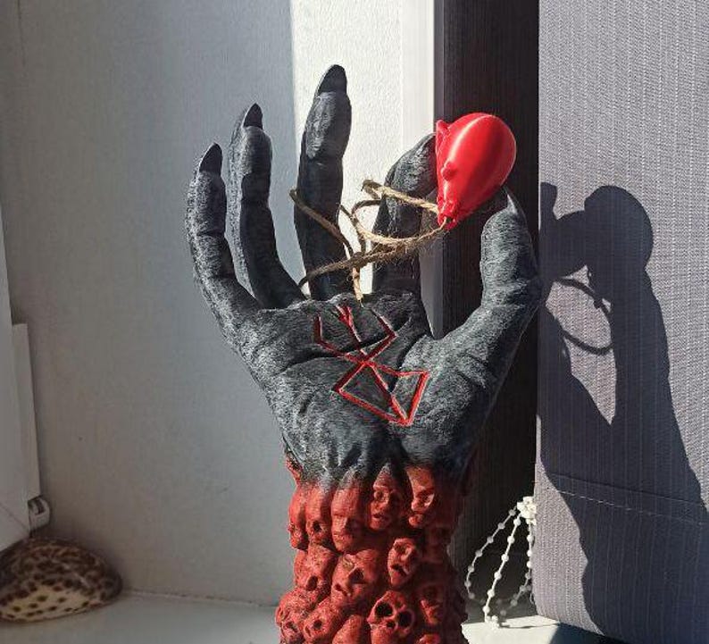 Puede incluir: Una escultura de mano negra con un s&iacute;mbolo rojo en la palma. La mano sostiene un objeto rojo con una cuerda atada. La mano est&aacute; hecha de muchas caras peque&ntilde;as.