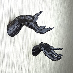 Puede incluir: Dos manos negras, impresas en 3D, de aspecto realista con largas y afiladas garras, montadas en una pared. Las manos están extendidas, con las palmas hacia adelante.