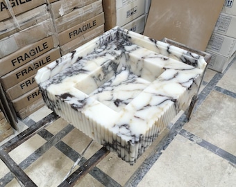 Può includere: Un lavandino in marmo bianco con venature nere. Il lavandino è rettangolare e ha un design unico e stratificato. È posizionato su un supporto in metallo.