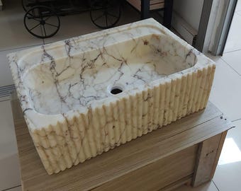Può includere: Un lavandino in marmo bianco con un bordo ondulato, posto su un piano di lavoro in legno. Il lavandino ha un foro di scarico e una finitura in marmo bianco.