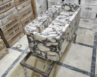 Può includere: Un lavabo rettangolare in marmo con un motivo venato bianco e grigio scuro. Il lavabo è sostenuto da un supporto in metallo scuro. Scatole di cartone con l'etichetta "FRAGILE" sono impilate sullo sfondo.