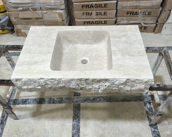 May include: A white stone bathroom sink with a rough, textured surface. The sink has a square shape with rounded edges and a rectangular basin. The sink is sitting on a tiled floor with black grout.