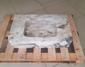 Puede incluir: Lavabo rectangular de mármol con un orificio de desagüe central. El lavabo presenta una base blanca con vetas marrones y negras. Está colocado sobre un palet de madera.