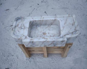 May include: A white marble sink with brown veining, mounted on a wooden stand. The sink is rectangular with rounded edges.