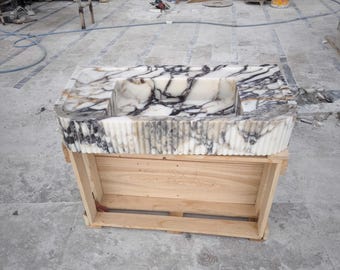 Puede incluir: Un lavabo de mármol blanco con vetas negras y doradas, colocado en una caja de madera. El lavabo tiene un borde ondulado y una forma rectangular.