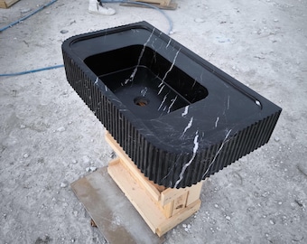 May include: A black marble sink with white veining, featuring a rippled edge and a rounded rectangular shape. The sink is resting on a wooden crate.
