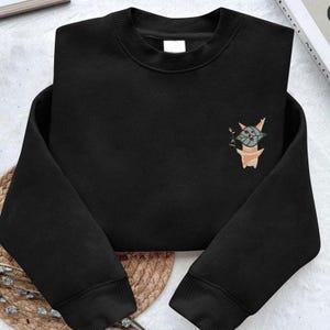May include: Black crewneck sweatshirt with an embroidered cartoon character design on the chest. The design features a light green and gray creature holding a small stick. The sweatshirt is folded neatly.