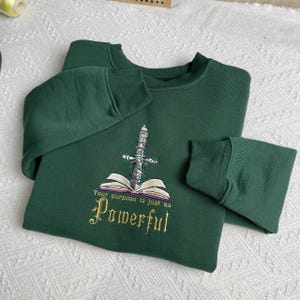 Może przedstawiać: Zielona bluza z haftowanym wzorem przedstawiającym miecz przebijający książkę. Pod wzorem znajduje się haft z tekstem "Your purpose is just as Powerful".