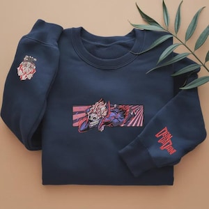 May include: Navy blue crew neck sweatshirt. The front of the sweatshirt displays a graphic of a skull with red and white hair against a red and pink backdrop. The left sleeve has a bulldog graphic, and the right sleeve has red text reading "Zan Tatan".