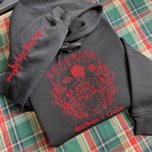 May include: Black hooded sweatshirt with a red embroidered design featuring a character with a sun and clouds, the text "Astarion Approves" and "Baldur's Gate".