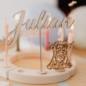 Könnte beinhalten: Ein Kuchenaufsatz aus Holz mit dem Namen "Julian" in Schreibschrift. Ein Highland-Kuh aus Holz und Kerzen umgeben den Namen. Der Kuchenaufsatz steht auf einer weißen Holzbasis mit einem hellorangenen Zentrum.