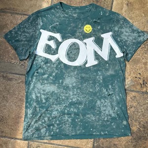 May include: A green t-shirt with a distressed, faded look. The shirt has a white graphic of the word "EOM" with a yellow smiley face above it.