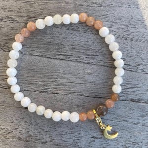 Könnte beinhalten: Ein weißes und pfirsichfarbenes Perlenarmband mit einem goldenen Halbmond-Anhänger.