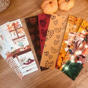 Peut inclure: Une collection de marque-pages colorés avec des illustrations sur le thème de l'automne. Les motifs comprennent des cœurs, des imprimés léopard et des scènes chaleureuses avec des citrouilles et le mot "Autumn". Les marque-pages sont disposés sur une surface en bois.