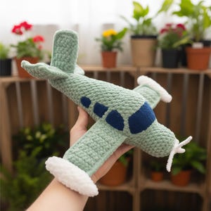 Puede incluir: Un avión de juguete tejido a mano en verde y azul. El cuerpo es verde claro con ventanas y detalles en azul oscuro. Las alas tienen ribetes blancos y la hélice es blanca. El juguete está diseñado para parecerse a un avión antiguo.