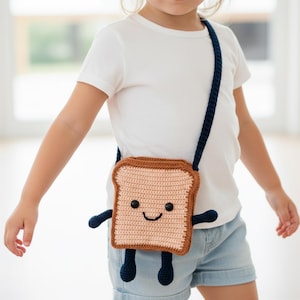 Op de afbeelding: Een gehaakte crossbodytas in de vorm van een toast met een lachend gezicht, zwarte ogen en armen en benen. De tas heeft een bruine korst en een beige midden, met een marineblauwe band. Gedragen door een kind.