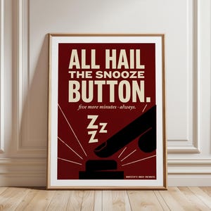 Könnte beinhalten: Gerahmtes Poster mit burgunderrotem Hintergrund und goldenem Rahmen. Der Text "ALL HAIL THE SNOOZE BUTTON" ist in großen, cremefarbenen Buchstaben dargestellt. Darunter steht der Text "five more minutes - always". Eine schwarze Illustration zeigt einen Finger, der auf einen Knopf drückt.