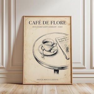 May include: A framed vintage-style print featuring a cafe scene. The artwork depicts a table with a cup of coffee, a newspaper, and the text "CAFÉ DE FLORE" and "BOULEVARD SAINT-GERMAIN - PARIS". The print has a sepia tone.