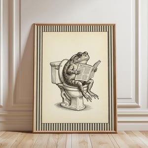 May include: A framed wall art print featuring a black and white ink-style illustration of a toad sitting on a toilet while reading a newspaper titled The Daily Pond. The artwork is set against a cream background with a black and white striped border.