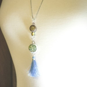 Puede incluir: Un collar de cadena plateada con un borla azul, una cuenta redonda con estampado verde y blanco y una cuenta redonda marr&oacute;n y dorada.