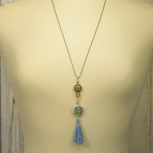 Puede incluir: Un collar largo de cadena plateada con un borla azul y tres colgantes redondos. Los colgantes est&aacute;n decorados con patrones verdes, marrones y dorados.