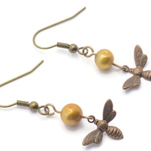 May include: A pair of gold-toned earrings featuring a small bee charm and a golden pearl bead on each earring.