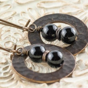 Peut inclure: Une paire de boucles d'oreilles pendantes avec des pendentifs ovales en bronze martel&eacute;, chacune avec deux perles noires. Les boucles d'oreilles ont de longs et fins fils d'oreille en bronze. Elles sont pr&eacute;sent&eacute;es sur une surface blanche textur&eacute;e.