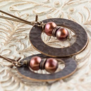 May include: A pair of handcrafted earrings featuring hammered copper oval and circular shapes. Each earring has two maroon-colored beads and long, thin copper wires for the ear. The earrings are displayed on a white, ornate surface.