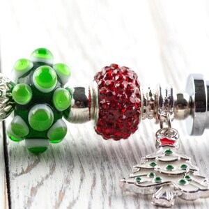 May include: A close-up of a charm bracelet with various beads and charms. The bracelet features a white faceted bead, a silver filigree bead, a green glass bead, and a red, textured bead. A silver Christmas tree charm hangs from the bracelet.