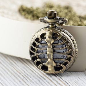 May include: A gold-toned pocket watch with a skeleton ribcage design. The watch face is visible through the ribs and has black numbers and hands.