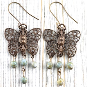 May include: A pair of bronze-colored butterfly earrings. The earrings feature intricate openwork wings and a central rectangular design. Each earring has three dangling, round, pale green and blue beads. The earrings hang from bronze-colored ear wires.