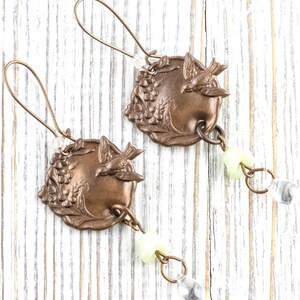 May include: A pair of bronze-colored earrings. Each earring features a circular pendant with a bird and grape design. The earrings have a long, curved hook and are adorned with a small, pale yellow bead and a clear, teardrop-shaped crystal.