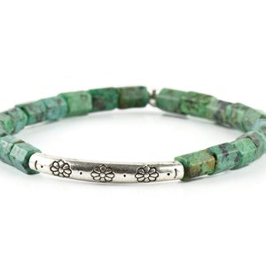 African Turquoise Stretch Bracelet on white background. Beads are bluish green with black and brown speckle. Silver pewter tube focal bead is adorned with floral etchings.